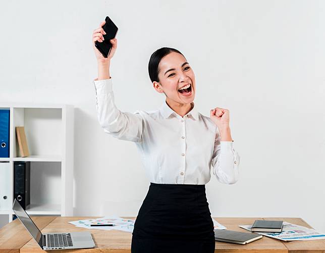 successful-young-businesswoman-holding-mobile-hand-clenching-her-fist-workplace