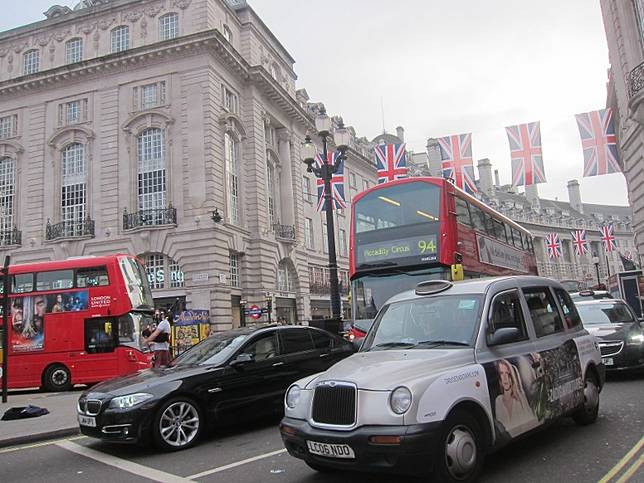 A busy city street decorated with Union Jack flags featuring a mix of traditional and electric vehicles, including a black car and a custom taxi, with classic architecture in the background and double-decker buses passing by.