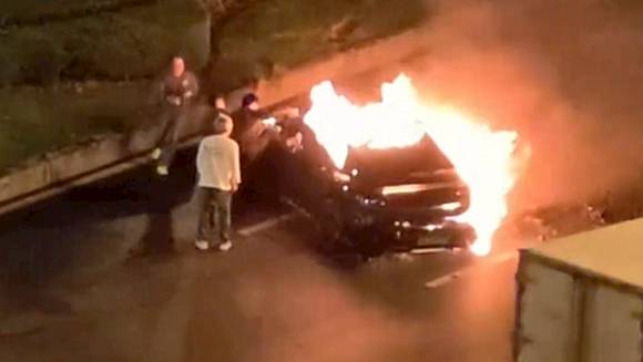 A black electric car, likely a Xiaomi model, is engulfed in flames on the side of a road with several individuals standing nearby, indicating an emergency response to a car fire possibly caused by a traffic accident involving drunk driving.