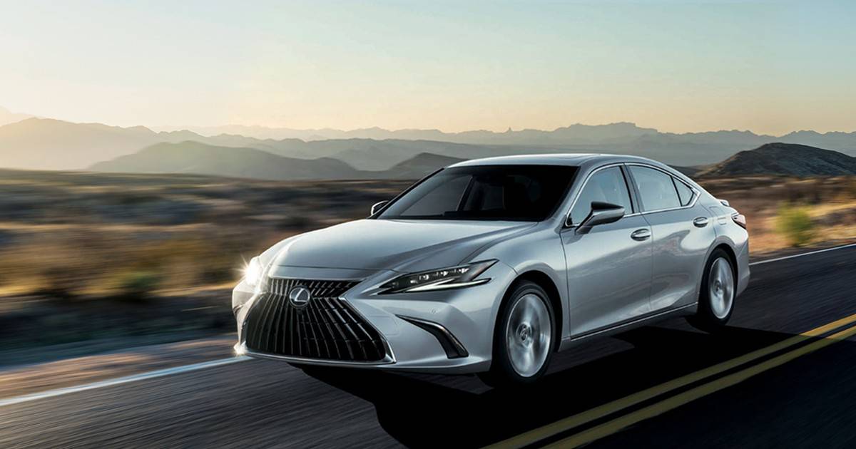 A silver Lexus ES hybrid sedan drives along an empty desert road during sunset, with distant mountain ranges in the background.