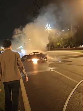A black electric vehicle is engulfed in flames on Tianfu Avenue in Chengdu, with smoke billowing into the air while emergency rescue efforts are presumably underway, and a man in a suit is observing the incident from the sidewalk.